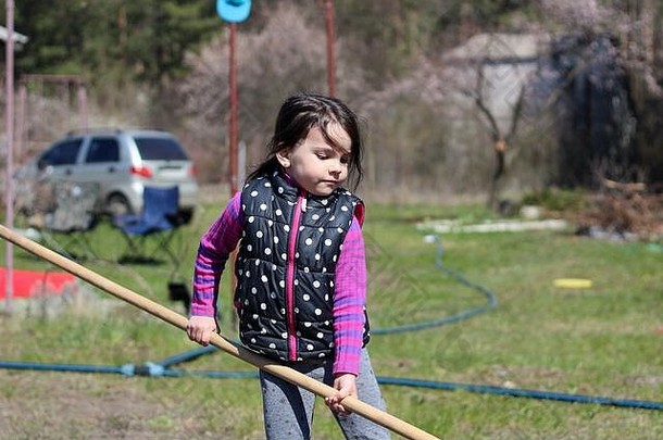 一个白色黑头发的白色小女孩，在户外扎着马尾辫，手里拿着一根长木棍。学龄前儿童在春天的花园里帮助父母