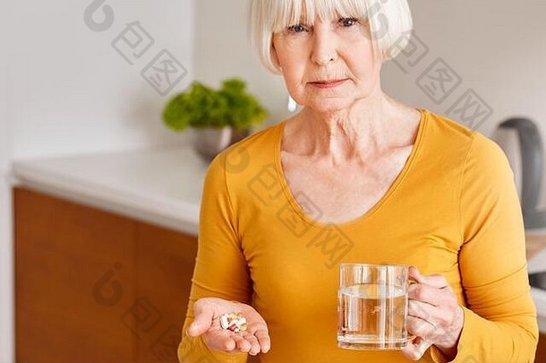 退休一名站在厨房的高级妇女拿着药片，在智能手机上搜索与冠状病毒有关的症状