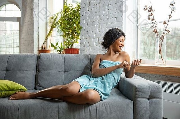 选美日。一位非洲裔美国妇女，穿着毛巾，准备在家里做日常护肤。坐在沙发上，修指甲，微笑。美容、自我护理、化妆品、青春、健康的理念。