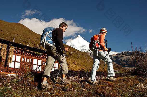 一对男女徒步经过一所房子，远处是吉楚德雷克山。他们两人正在不丹进行为期11天的长途跋涉。