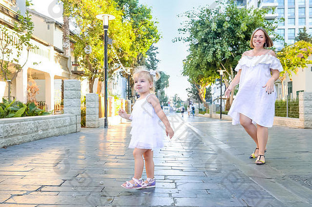 在城市公园散步时，可爱的情绪化的金发小女孩穿着衣服和怀孕的母亲玩耍，捕捉肥皂泡。活跃家庭