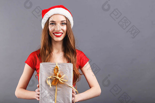 美丽的圣诞妇女戴着圣诞帽，穿着红色连衣裙，看着相机，送礼物，新年，圣诞节，节假日，纪念品，礼物，购物，折扣，商店，雪人圣诞老人，化妆，发型，狂欢节。