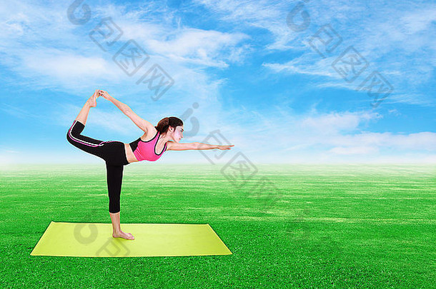 年轻女子在绿草蓝天上用瑜伽垫做瑜伽运动