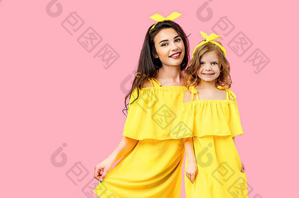 在粉色背景的摄影棚里，正面的黑发母亲抱着微笑的女儿，穿着同样的黄色衣服，看着摄像机