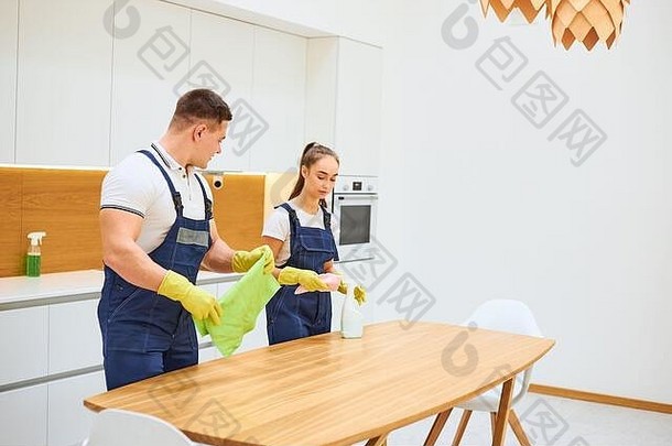 清洁服务的年轻白人工人一起在厨房里工作，擦去桌子上的灰尘。白色厨房