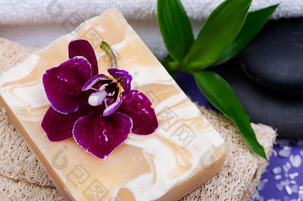 Spa健康概念。天然丝瓜海绵、杏仁山羊奶皂、白色毛巾、玄武岩石头、紫色背景上的竹子和兰花。
