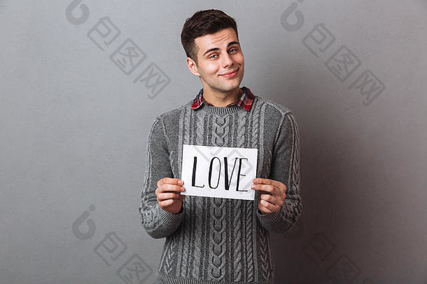 照片中，一个微笑的年轻人孤零零地站在灰色的墙上，手里拿着一张写着爱情文字的纸。看镜头。