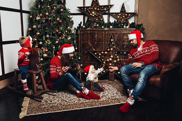穿着冬衣和红色圣诞帽的幸福家庭在圣诞树旁放松。