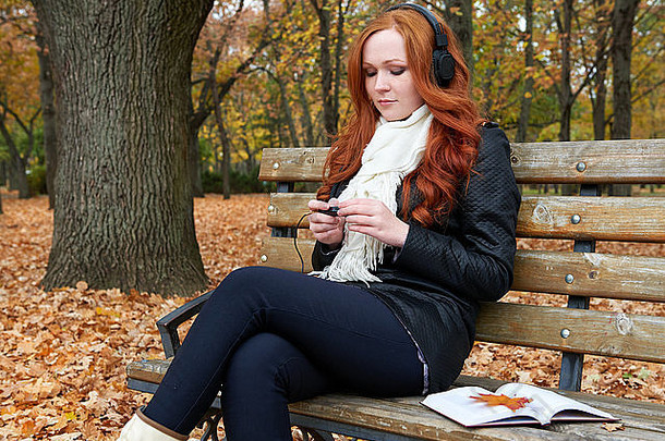 秋天，红发女孩在城市公园用耳机听音乐