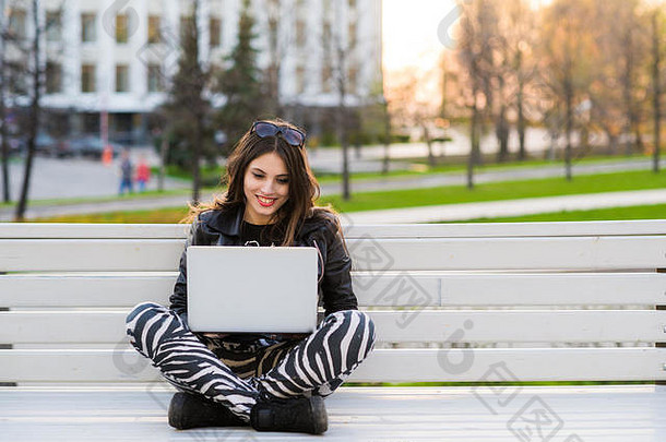 一名快乐的女学生坐在长凳上，手提电脑在户外，看着相机的照片