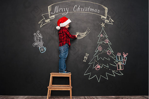 照片可爱的孩子穿他站凳子圣诞节树指出星星黑板