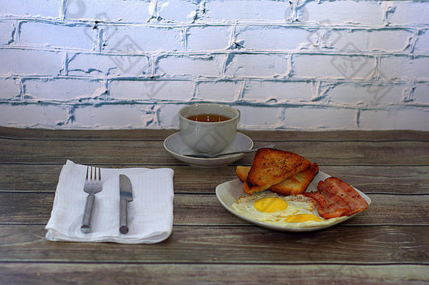 木桌上摆着一个白色的陶瓷盘子，上面放着煎蛋、熏肉和烤面包，旁边是一张餐巾纸，餐巾纸上有刀叉，还有一杯热调味品