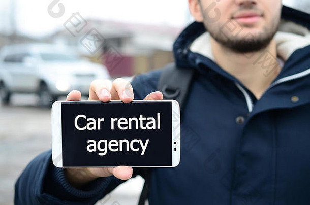 冬天，一个年轻人在一辆汽车和一幢大房子的背景上，在智能手机的显示屏上显示了一段文字。汽车租赁公司