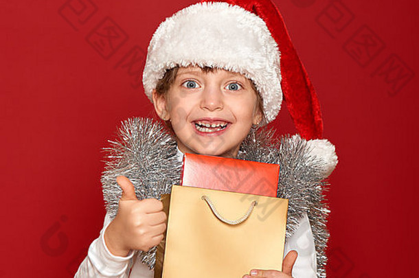 冬天，孩子，圣诞节概念-戴着圣诞帽的快乐女孩，红色背景上有一盒礼物