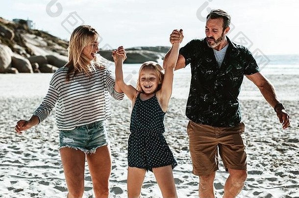 一对夫妇牵着女儿的手在海滩上玩耍。一个和父母一起度过暑假的女孩。