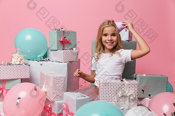 照片中，一个微笑的小女孩戴着生日帽，坐在一堆礼物盒里，看着粉色后背上的相机庆祝生日