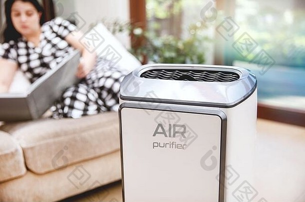 空气净化器净化空气。现代的空气净化器在客厅里净化空气，背景是一个坐在沙发上看书的女人。商标