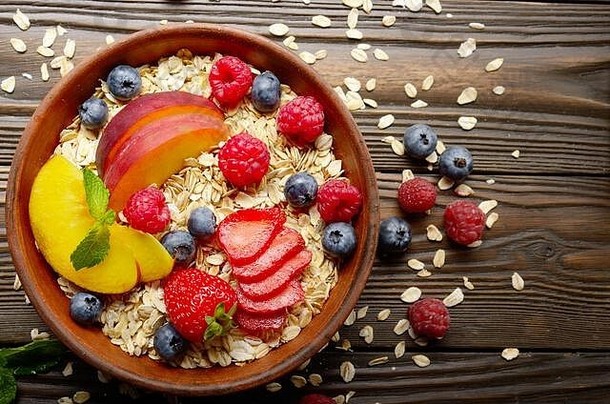 在木制厨房餐桌上的粘土盘中，平铺着水果健康的慕斯利，配以桃子、草莓、杏仁和黑莓
