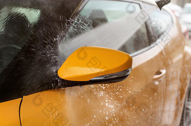 黄色汽车的侧后视镜在自助洗车机中清洗，喷射的水花击中车窗，水滴向四周飞扬。