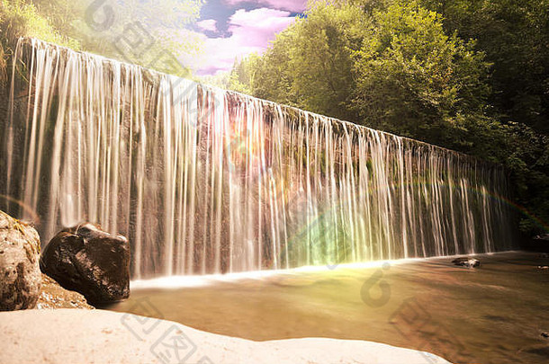 在风景如画的瀑布或瀑布中流过壁架或坝墙的水，绿叶葱郁，日落时阳光闪烁，彩虹闪烁