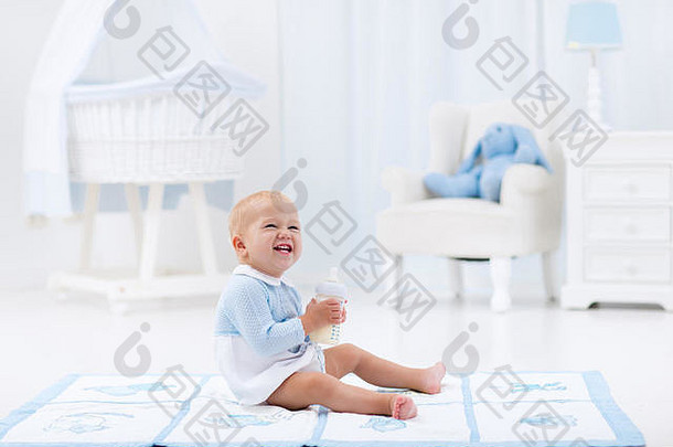 可爱的小男孩在一个有摇椅和摇篮的阳光明媚的白色托儿所里，在蓝色的地垫上玩耍，喝着奶瓶里的牛奶。卧室间