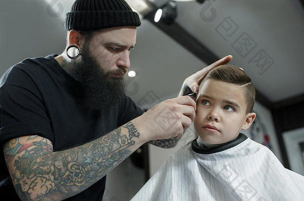 儿童理发师在黑暗的背景下给小男孩理发。心满意足的可爱学龄前男孩正在理发。大师的手上有“剃须”这个字的纹身