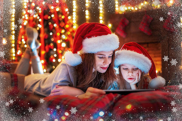 圣诞节时间。躺在床上，戴着圣诞老人帽、穿着睡衣、微笑着观看有趣视频或在数码平板电脑上挑选礼物的家庭母女