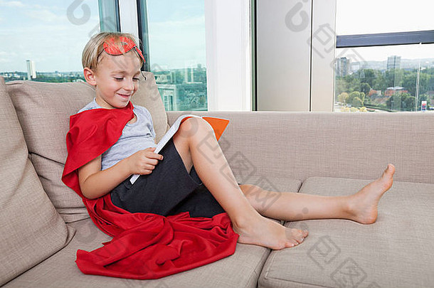 穿着超级英雄服装的微笑男孩在家的沙发上看书