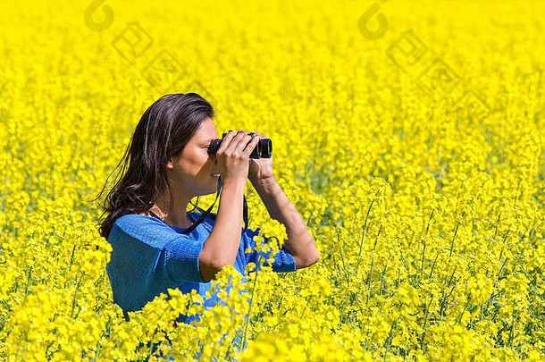 一位年轻女子用双筒望远镜在开花的黄色油菜地里观察