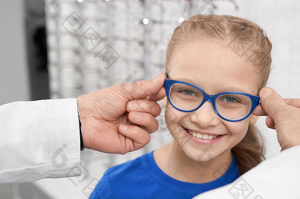 戴蓝色眼镜的孩子的特写镜头。男医生眼科医生帮助漂亮女孩选择眼镜。眼科诊所的医生和病人。保健的概念。