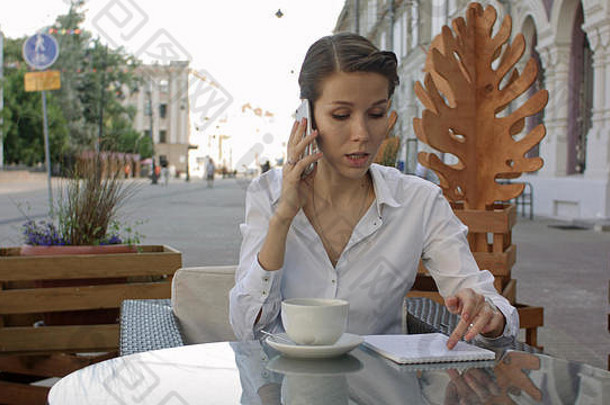 一张年轻的女商人坐在户外咖啡馆里放松地喝着咖啡，打着手机的照片