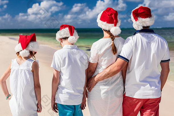 热带海滩上戴着红色圣诞老人帽庆祝圣诞节的美丽家庭的后视图