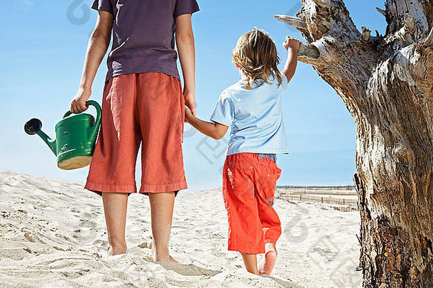 两个男孩（7-12岁）拿着水壶，站在海滩上的枯树旁