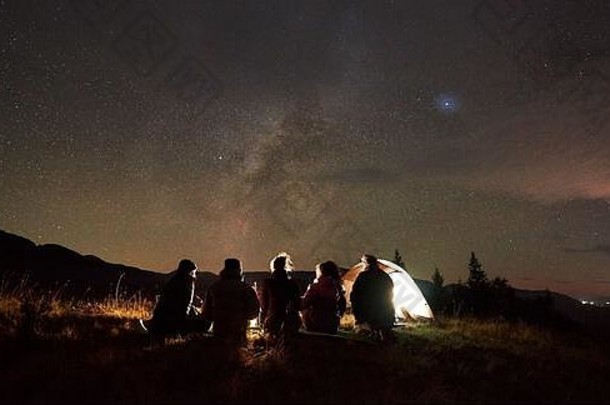 夜宿星空，旅游和积极的生活方式。五个人坐在游客帐篷的草谷上，在黑暗的星空背景下燃烧着营火。