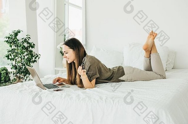 积极的<strong>女生</strong>躺在床上，用笔记本电脑。她正在屏幕上阅读，或者正在观看视频或培训视频课程，或者在线<strong>聊天</strong>。