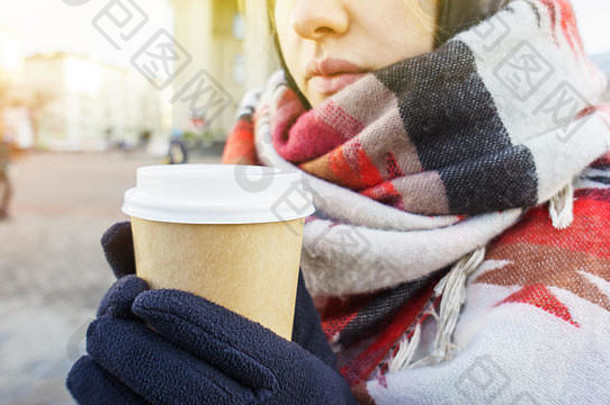 在城市环境中，戴着手套，戴着舒适的大围巾，手里拿着一杯外卖咖啡的年轻漂亮女孩。冬天喝咖啡的概念