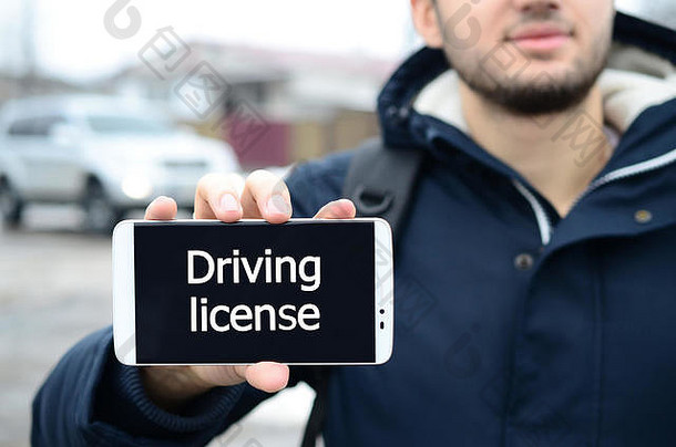 冬天，一个年轻人在一辆汽车和一幢大房子的背景上，在智能手机的显示屏上显示了一段文字。驾驶执照