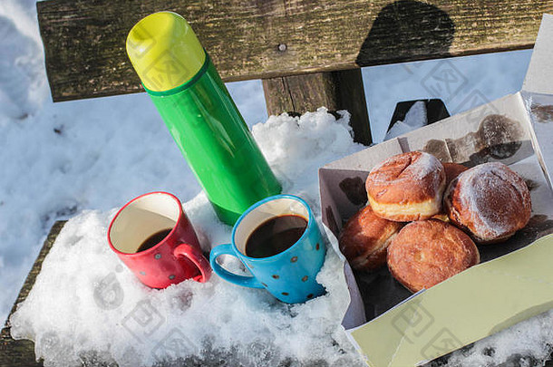 塞尔维亚贝尔格莱德附近的科斯马伊山上，在覆盖着积雪的木凳上，绿色的保温瓶、红色和蓝色的咖啡杯以及带甜甜圈的盒子