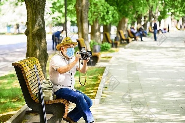 摄影师把相机放在长凳上。禁止参观公共场所。戴着医用面罩的老人。老人在公园里休息。冠状病毒锁定期间的生命。享受美丽。对疾病的恐惧。