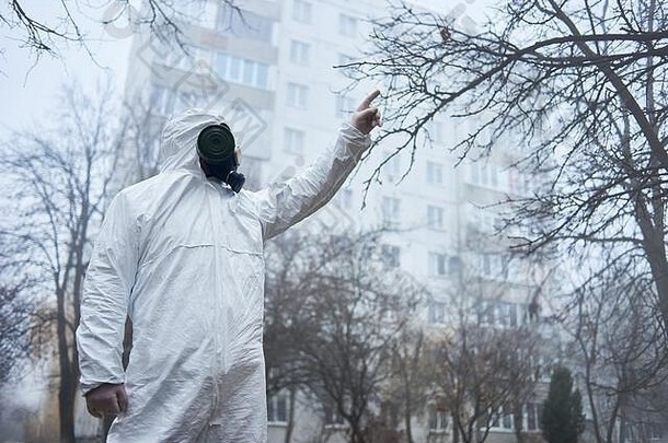 科学家站在秋天的树木之间，用手指指着天空。戴着防毒面具和防护服的男子在建筑物附近的空城里摆姿势。<strong>城市污染</strong>、恶劣环境、雾的概念。