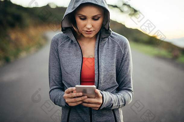 一位穿着连帽运动衫走在街上的健身女士看着她的手机。一名妇女在一条空荡荡的路上用手机行走。