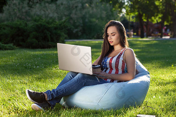 年轻的可爱的女孩移动PC坐豆袋花园公园绿色草在线购物概念