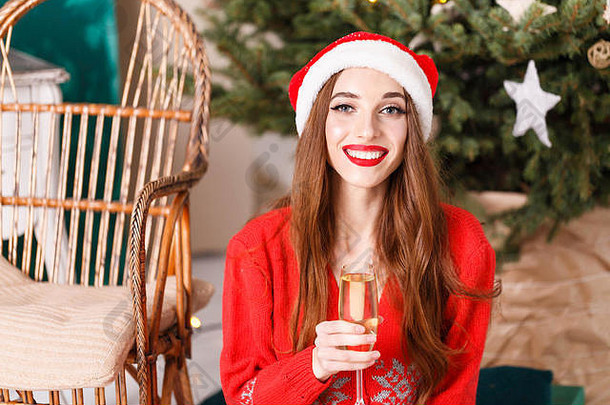 穿着圣诞帽和红色毛衣的迷人圣诞女郎坐在舒适的新年云杉旁的地板上，喝着香槟、新年、圣诞节、假日、纪念品、礼物、购物、折扣、商店、雪人圣诞老人、化妆、发型、狂欢节。