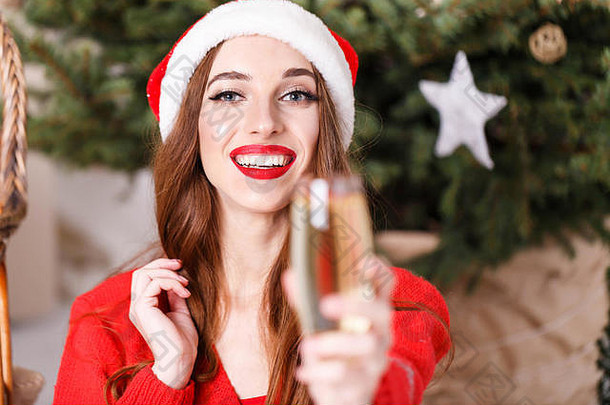 穿着圣诞帽和红色毛衣的迷人圣诞女士坐在舒适的新年云杉旁的地板上，一边说祝酒词，一边喝香槟，新年、圣诞节、假日、纪念品、礼物、<strong>购</strong>物、折扣、商店、雪人圣诞老人、化妆、发型、<strong>狂欢</strong>节。