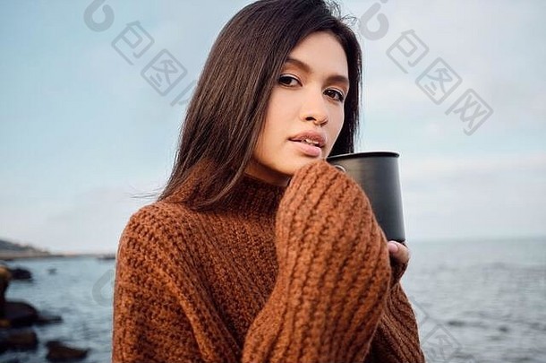 美丽的亚洲女孩，穿着针织毛衣，手里拿着杯子，在海边凝视着镜头