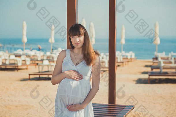 真正可爱的孕妇，穿着白色轻快的连衣裙，行走在沙滩上，用木制调色板搭桥，抱着肚子。美丽迷人的年轻女孩e