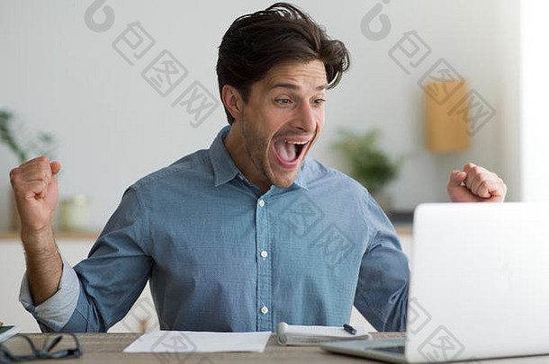 在笔记本电脑上庆祝成功的家伙在工作场所挥舞着拳头