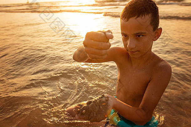 深思熟虑的男孩坐着海海岸日落玩沙子