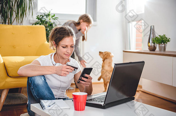一名身穿白衬衫、戴着耳机的妇女在智能手机上网上购物，并使用，背景是一个女儿和一条狗