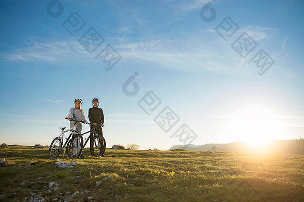 一对骑着山地自行车的夫妇站在夜空<strong>下</strong>的小山上，享受着日落时的灿烂阳光。
